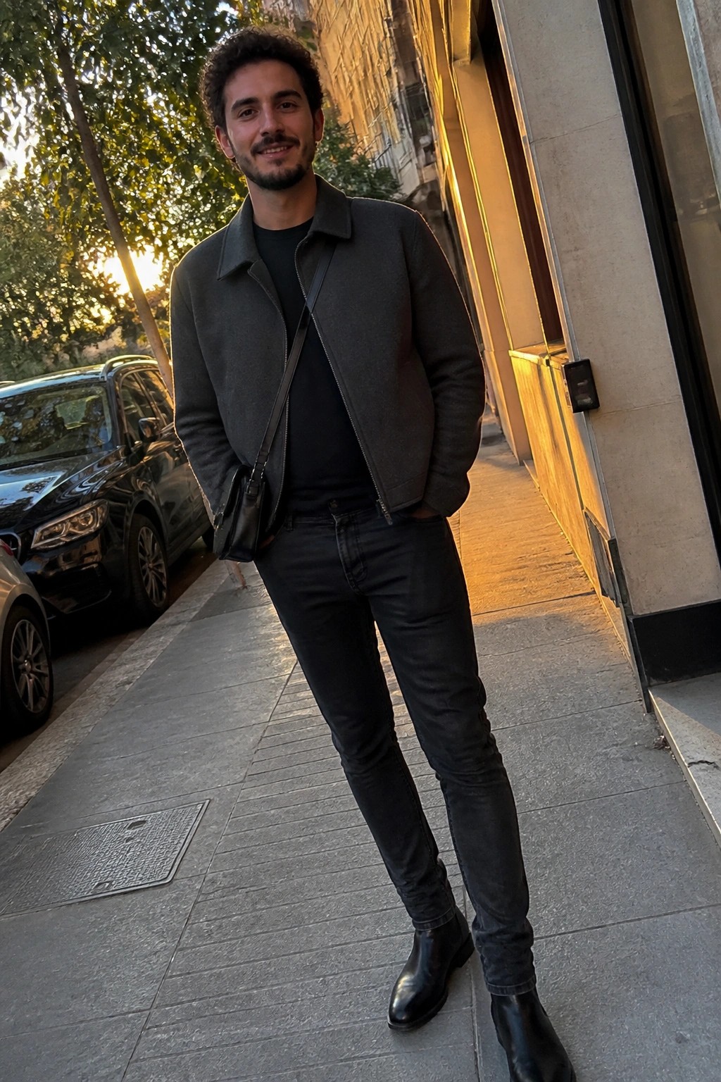 Curly-haired man in gray jacket and black jeans on sunlit city sidewalk