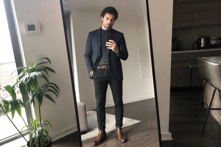 21 Smart Men’s Fall Semi Formal Outfits for Evening Dinner and Date Nights Man in navy blazer over gray sweater with black pants brown belt and brown Chelsea boots taking a mirror selfie in a modern apartment.