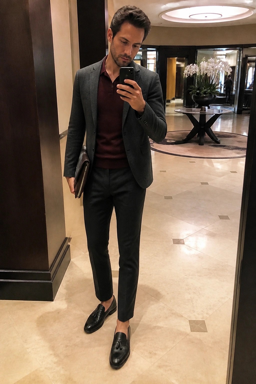 Man in gray blazer over maroon sweater taking mirror selfie in elegant lobby