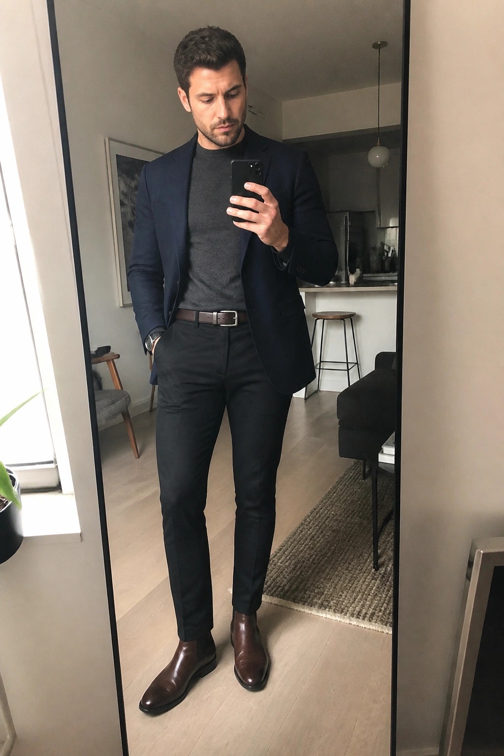 Man in navy blazer over gray sweater with black pants brown belt and brown Chelsea boots taking a mirror selfie in a modern apartment.
