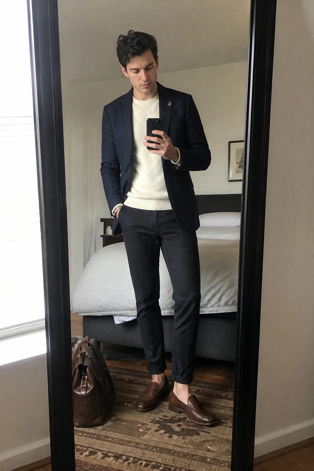 Man in navy blazer over white sweater checks phone in bedroom mirror