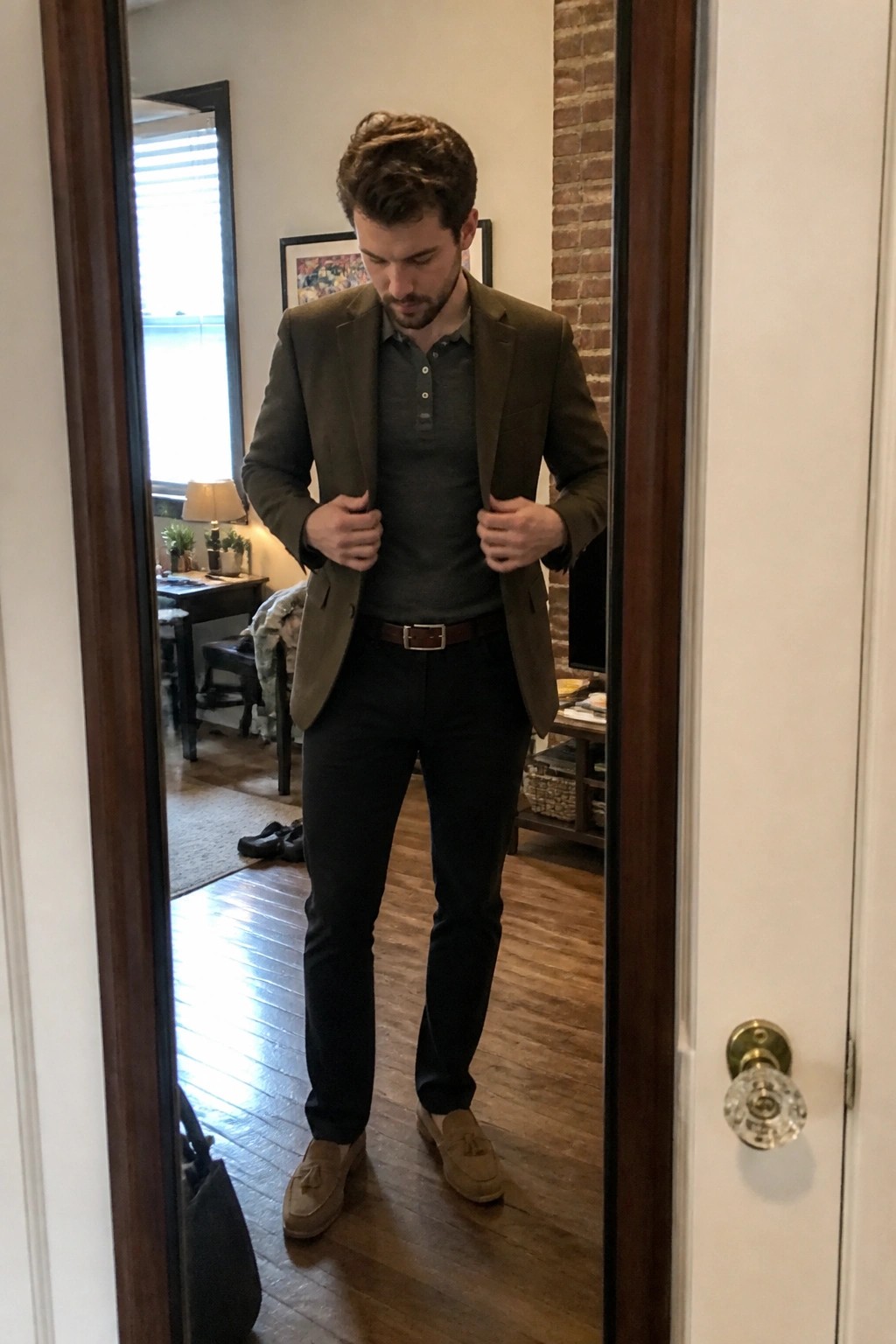 Man adjusting olive blazer over gray polo in mirror, black pants and brown shoes