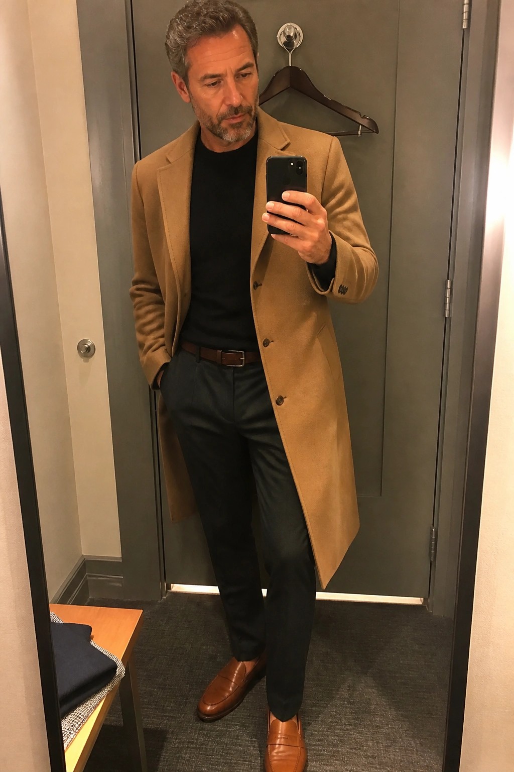 Man in tan overcoat and black sweater taking mirror selfie with phone