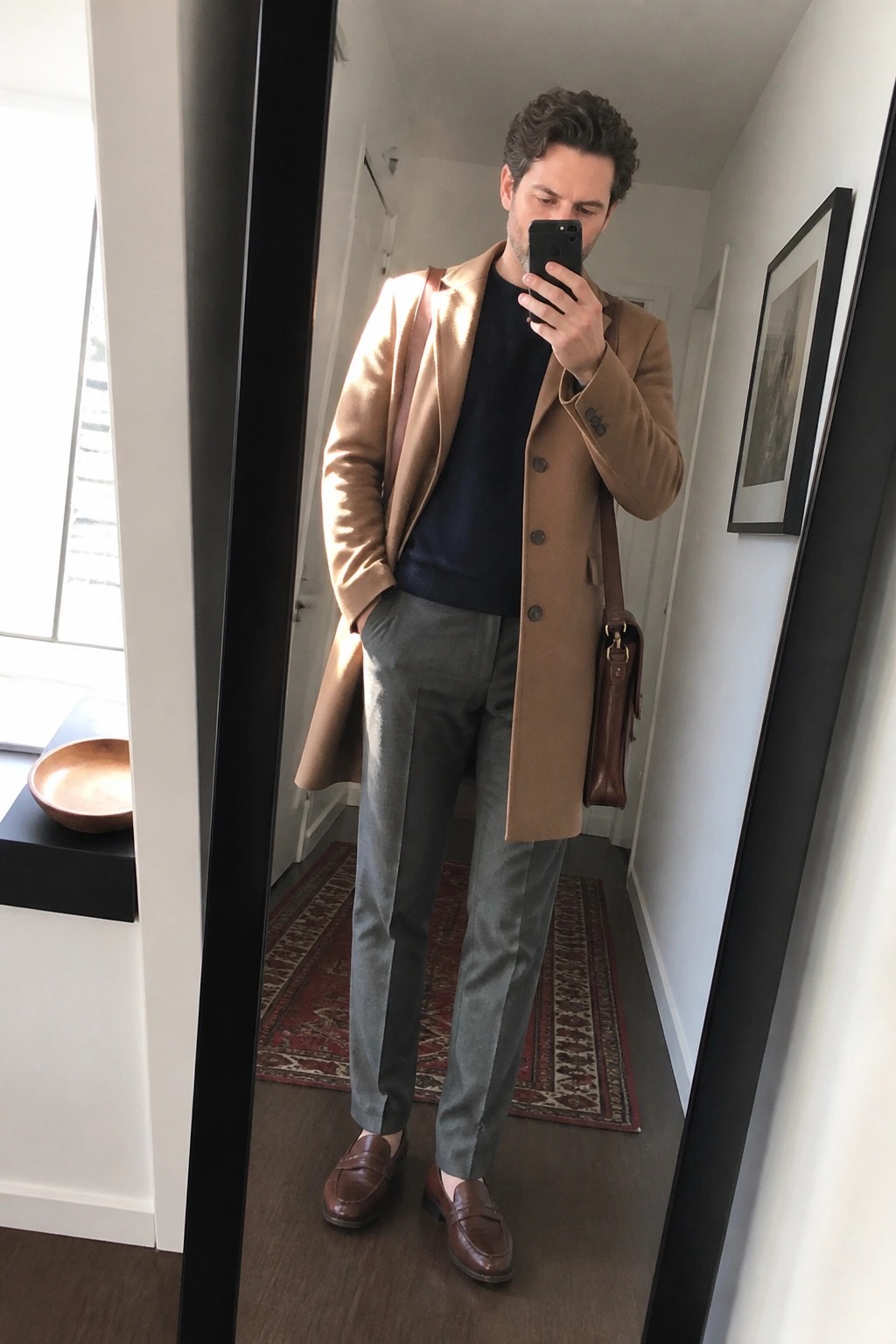 Man in tan overcoat over navy sweater taking mirror selfie in hallway