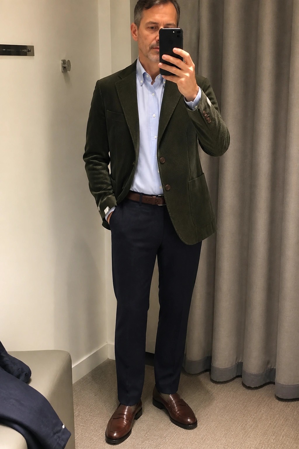Man in green corduroy blazer taking mirror selfie in blue shirt and navy pants