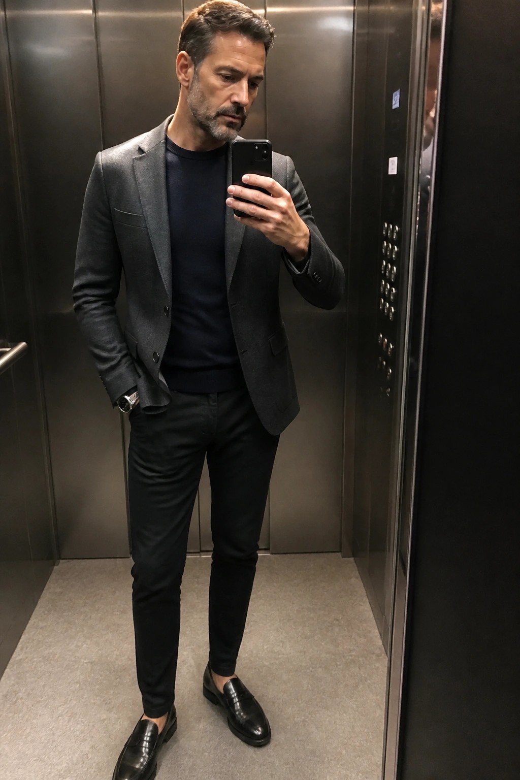 Man in gray blazer and navy sweater taking phone selfie in elevator