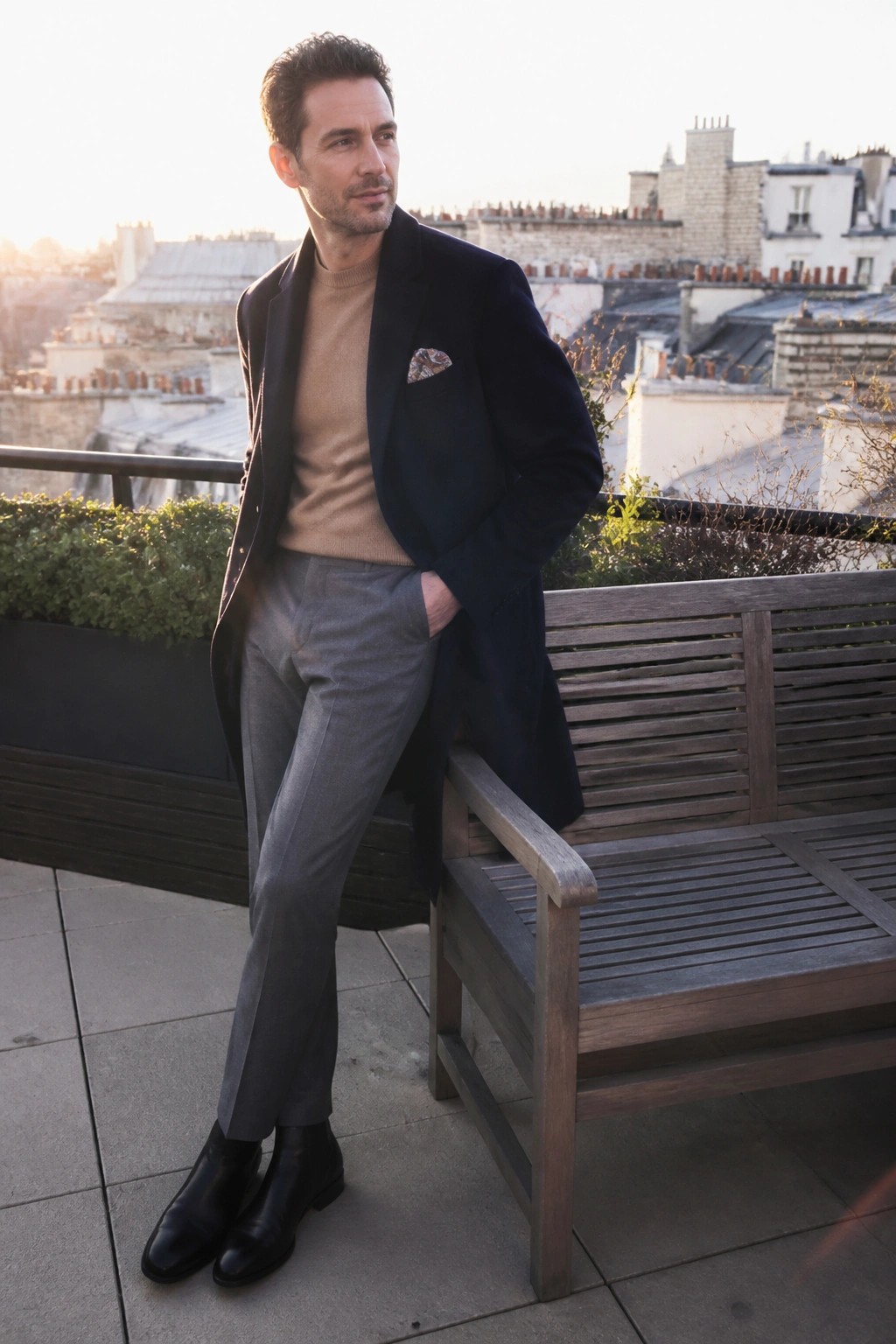 Man in navy overcoat over beige sweater and gray trousers on rooftop terrace at sunset