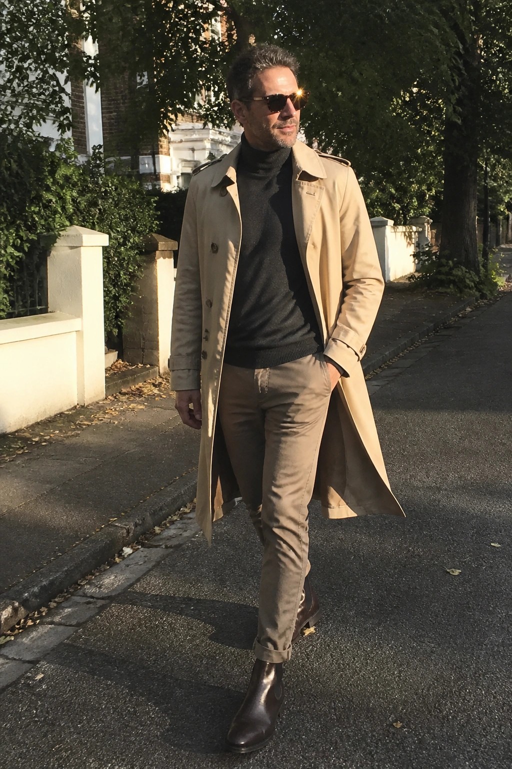 Man in beige trench coat over black turtleneck walking on sunny sidewalk
