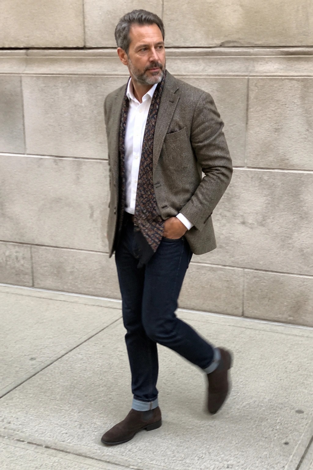 Man in tweed blazer, patterned scarf, jeans, and suede boots by stone wall
