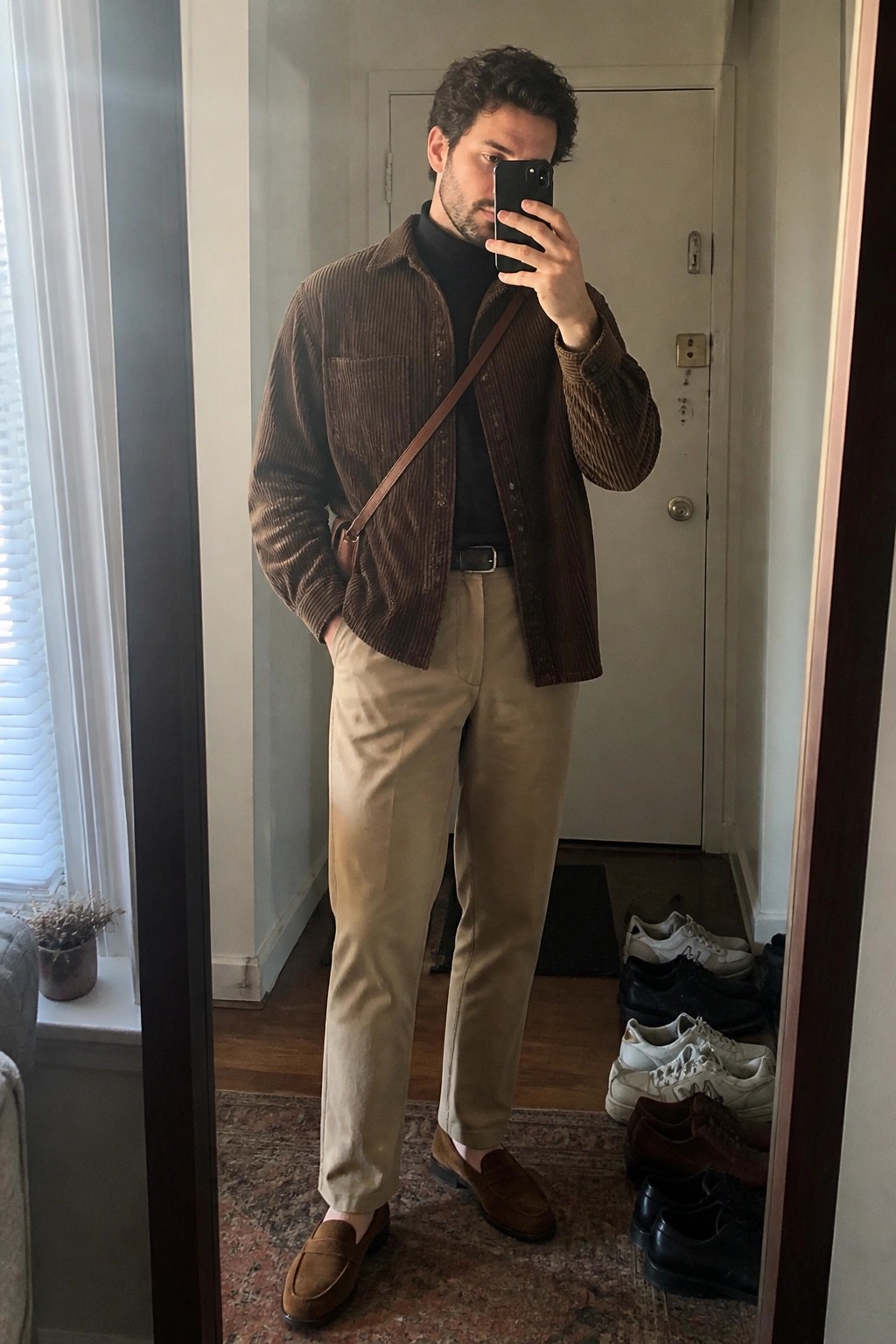 Man in brown corduroy shirt over black turtleneck taking mirror selfie in beige pants.