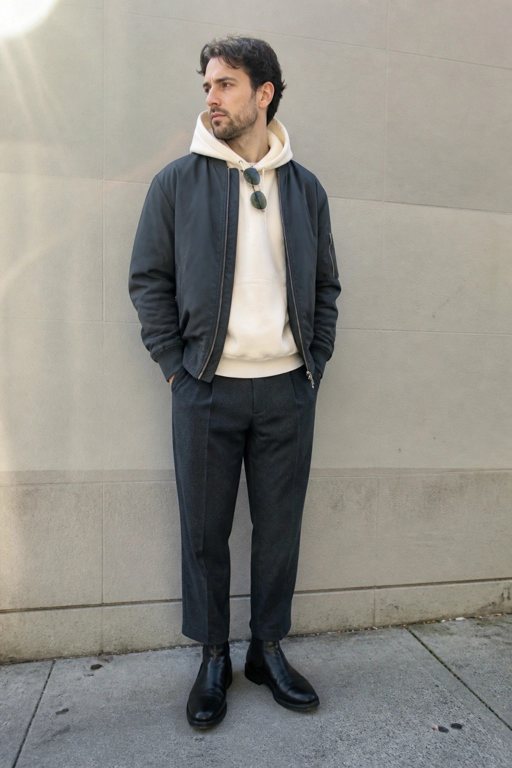 Man in dark bomber jacket over cream hoodie with sunglasses, gray pants, black boots.