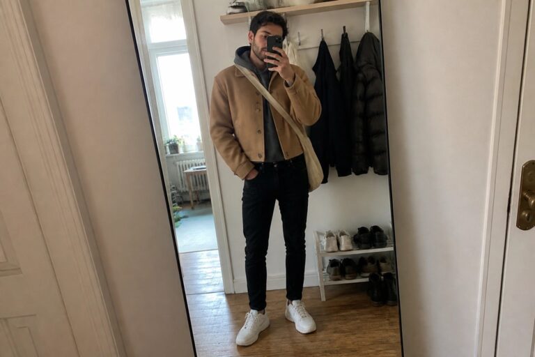 Man with curly hair in tan jacket taking mirror selfie with beige crossbody bag