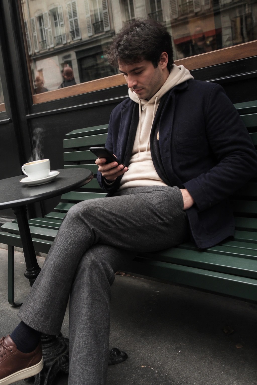 Man in navy jacket over beige hoodie checks phone on green bench beside coffee.