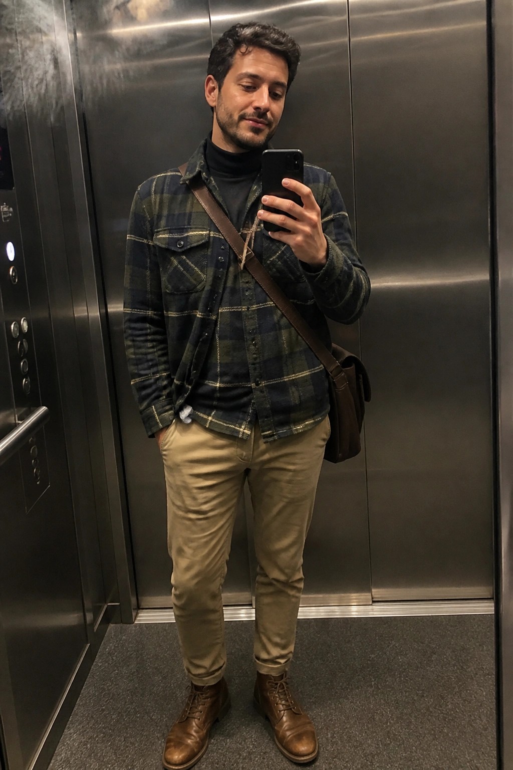 Man in plaid shirt and khaki pants taking selfie in metal elevator with bag