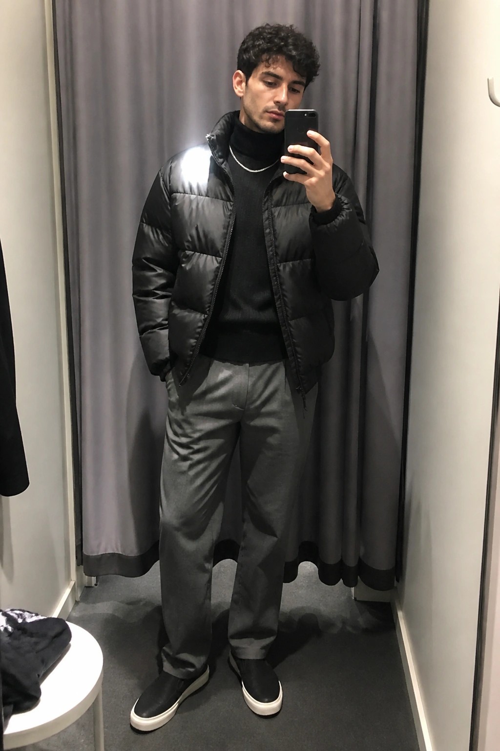 Man in black puffer jacket over black turtleneck with silver chain necklace, gray trousers, and black slip-on sneakers taking a mirror selfie in a fitting room.