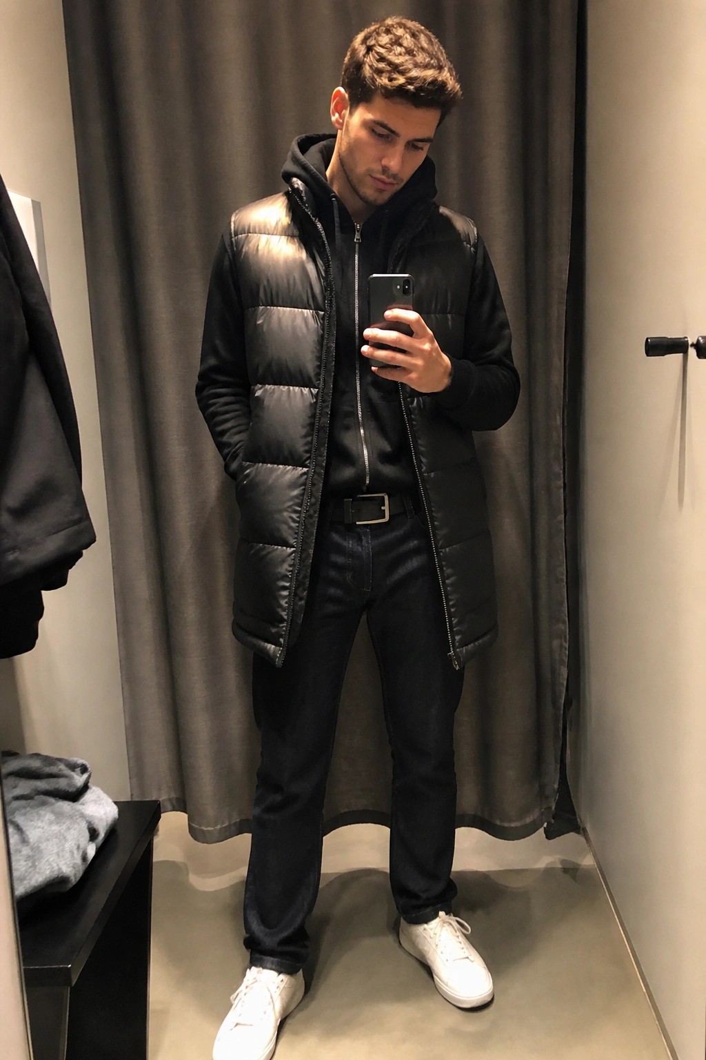 Man in black puffer vest checking smartphone in gray-curtained fitting room mirror