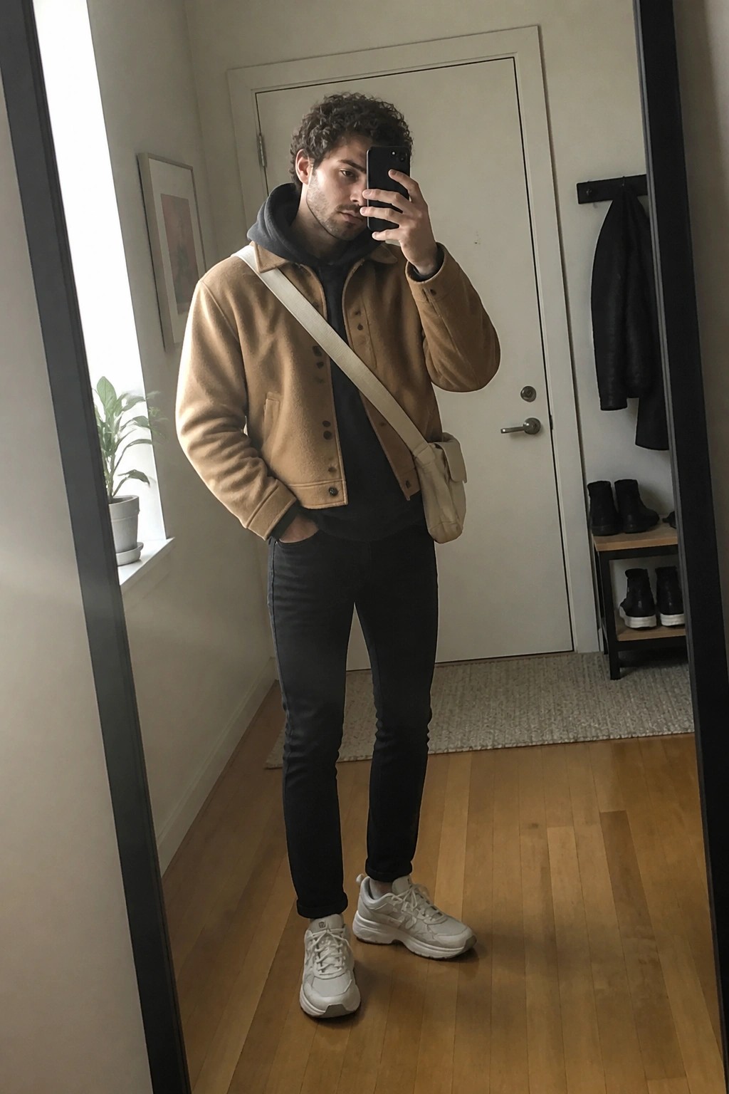 Man with curly hair in tan jacket taking mirror selfie with beige crossbody bag