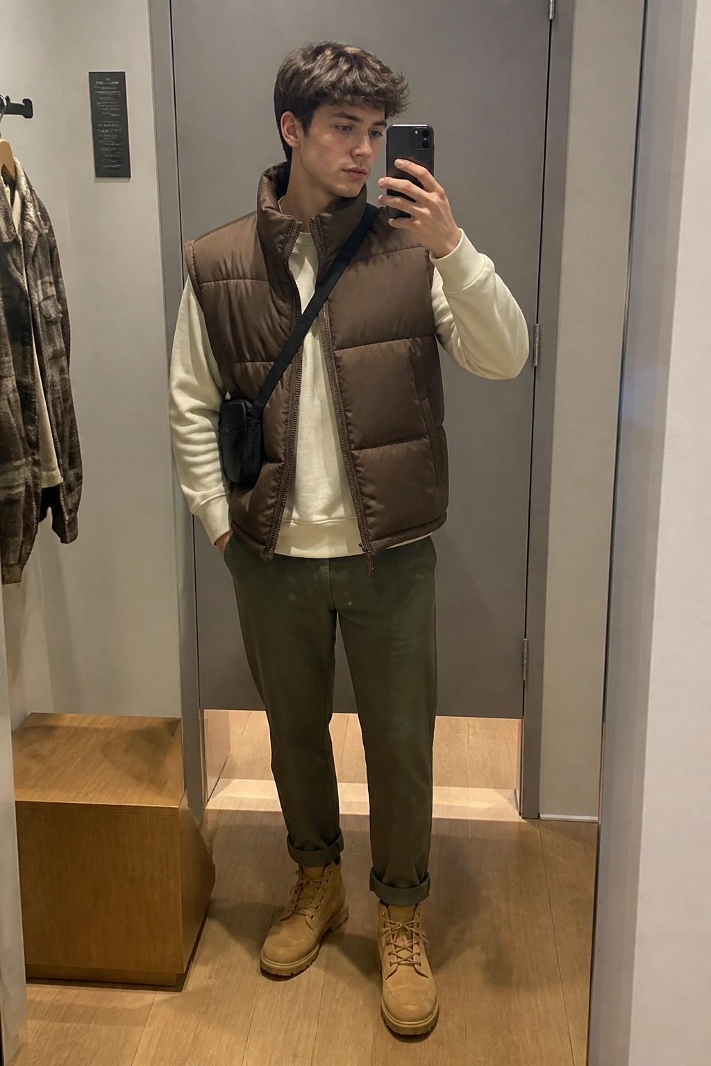 Young man in brown puffer vest, white sweatshirt, green pants, and tan boots takes mirror selfie.