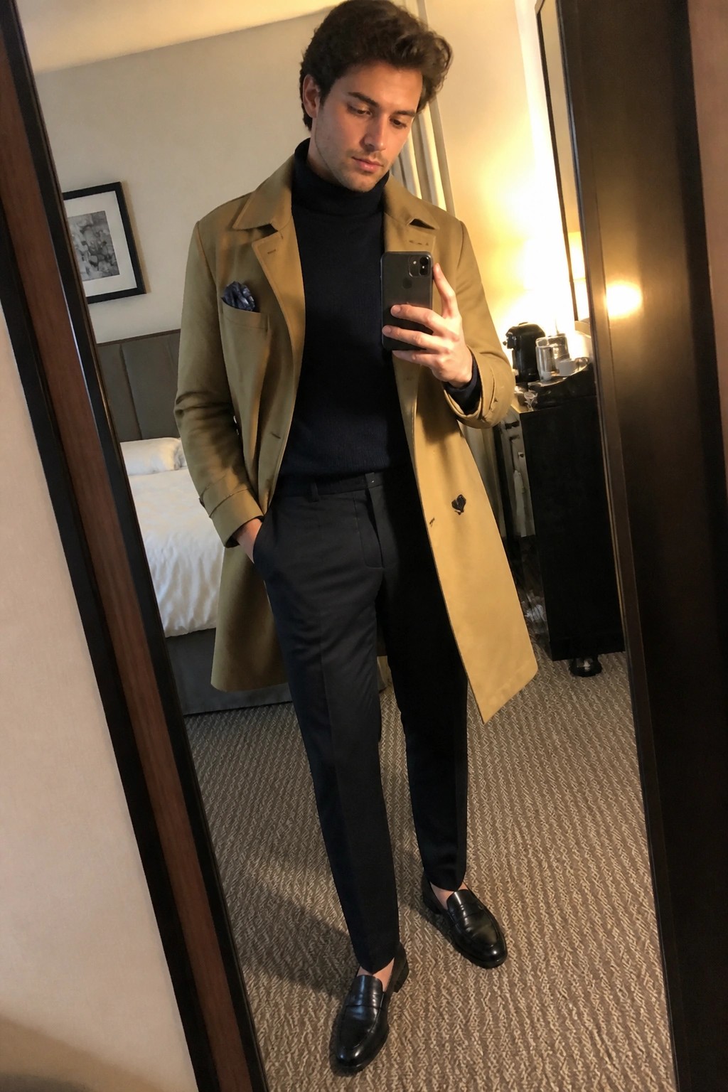 Man in tan overcoat over black turtleneck taking mirror selfie in hotel room