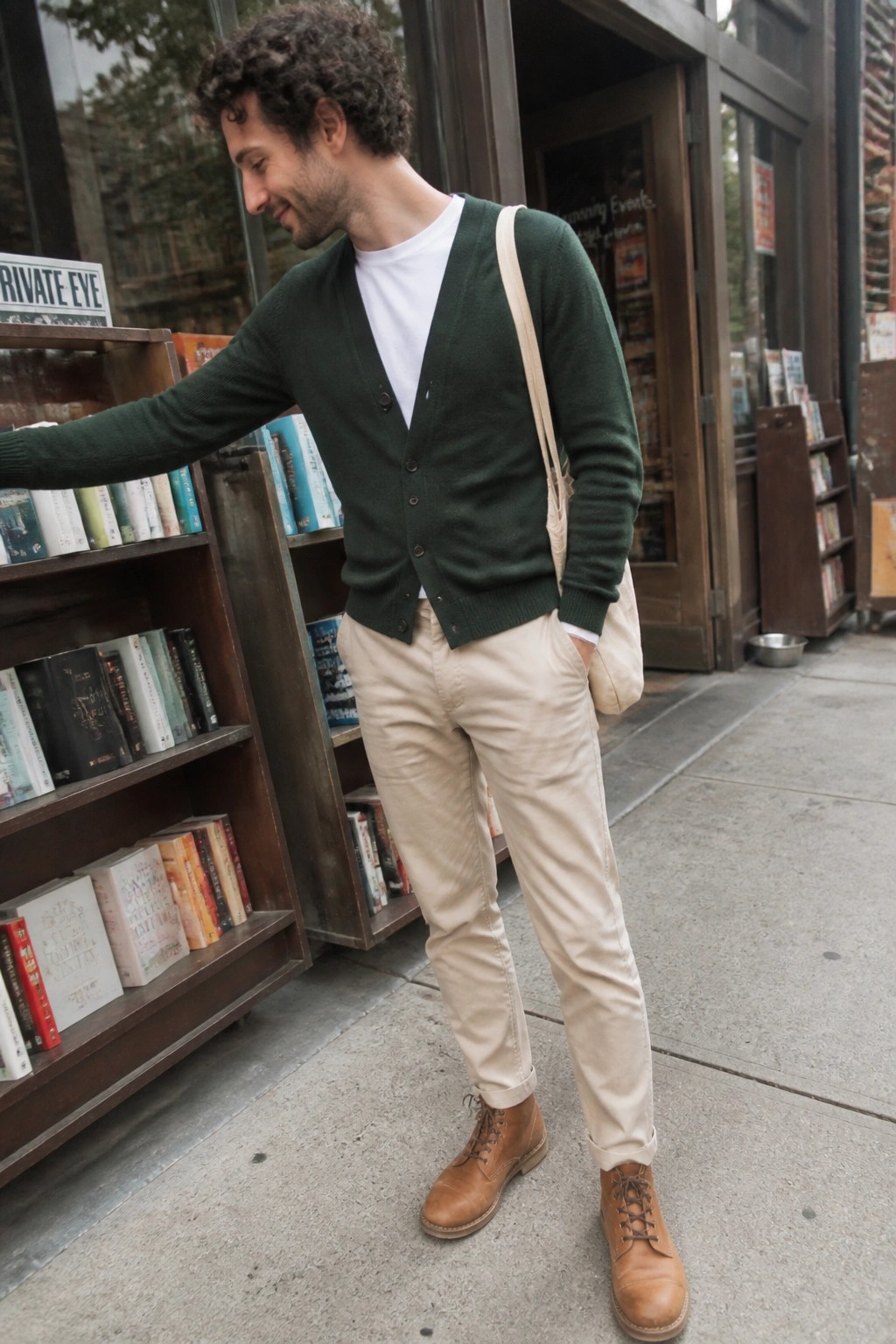 Man in green cardigan reaching for books on outdoor sidewalk shelf.