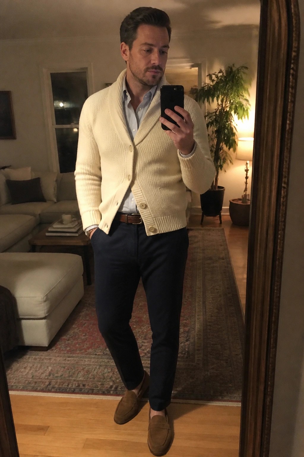 Man in cream cardigan and navy pants taking mirror selfie in living room