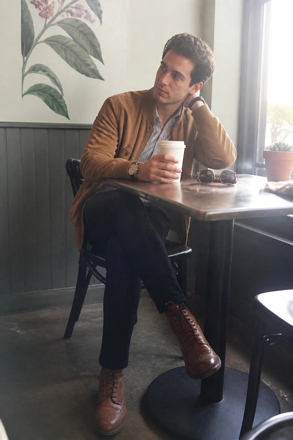 Man in brown suede jacket sits casually at cafe table holding coffee