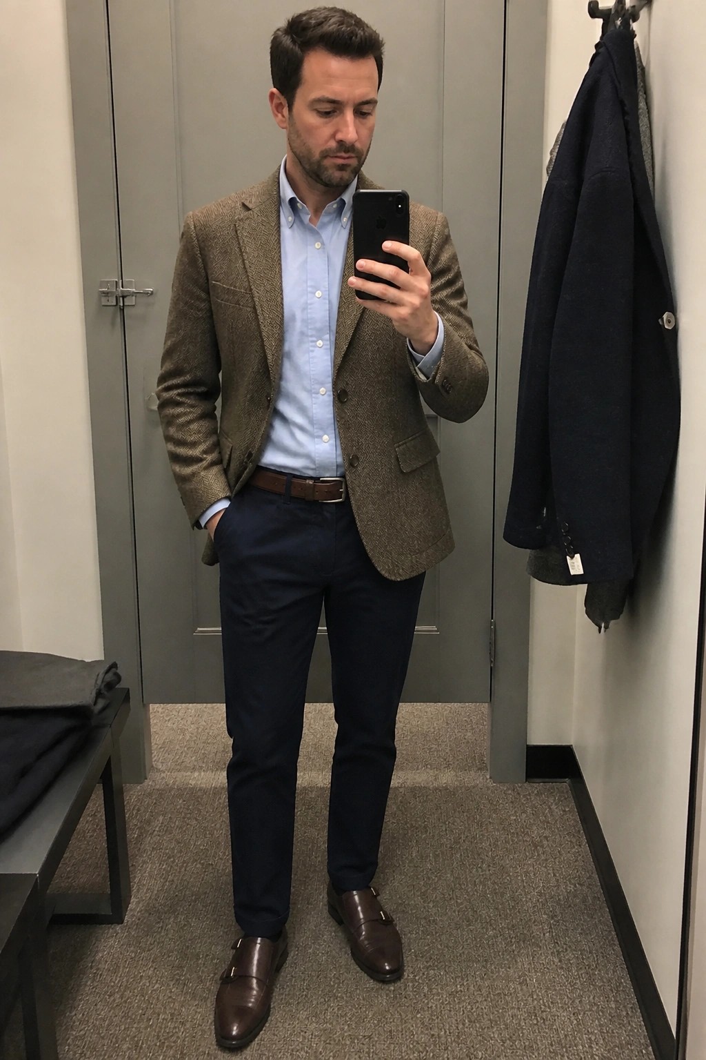 Man in brown herringbone blazer, blue shirt, and navy pants taking mirror selfie
