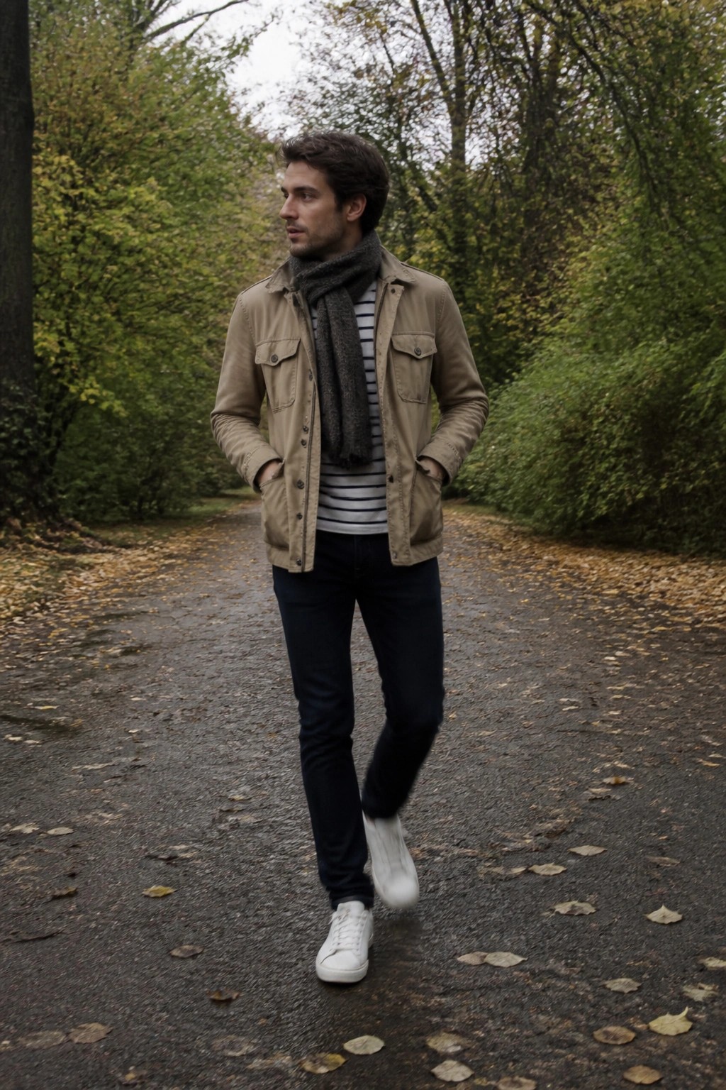 Man in beige jacket, striped shirt, gray scarf walks on autumn leaf path.