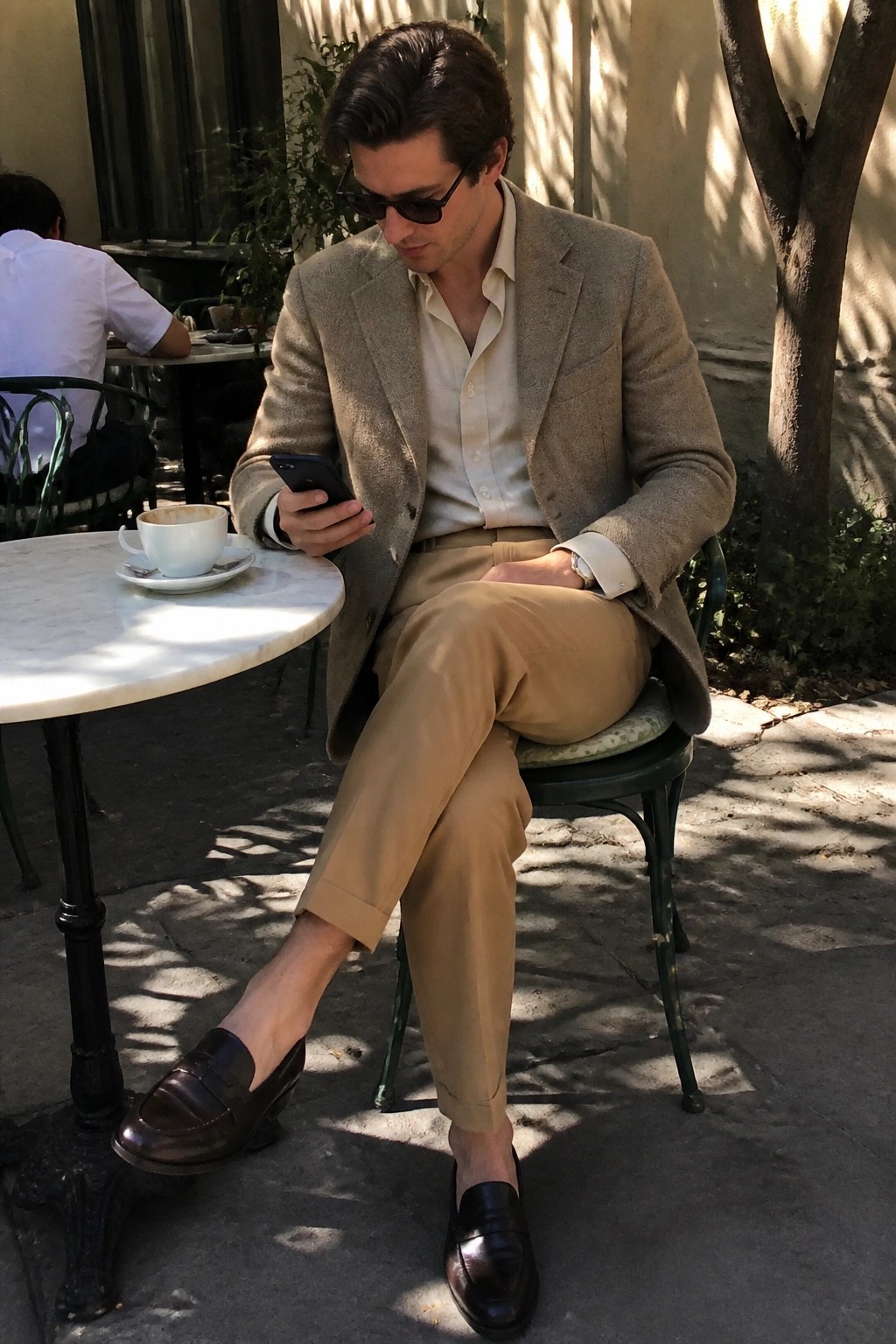 Man in beige blazer and tan pants checks smartphone at outdoor cafe table