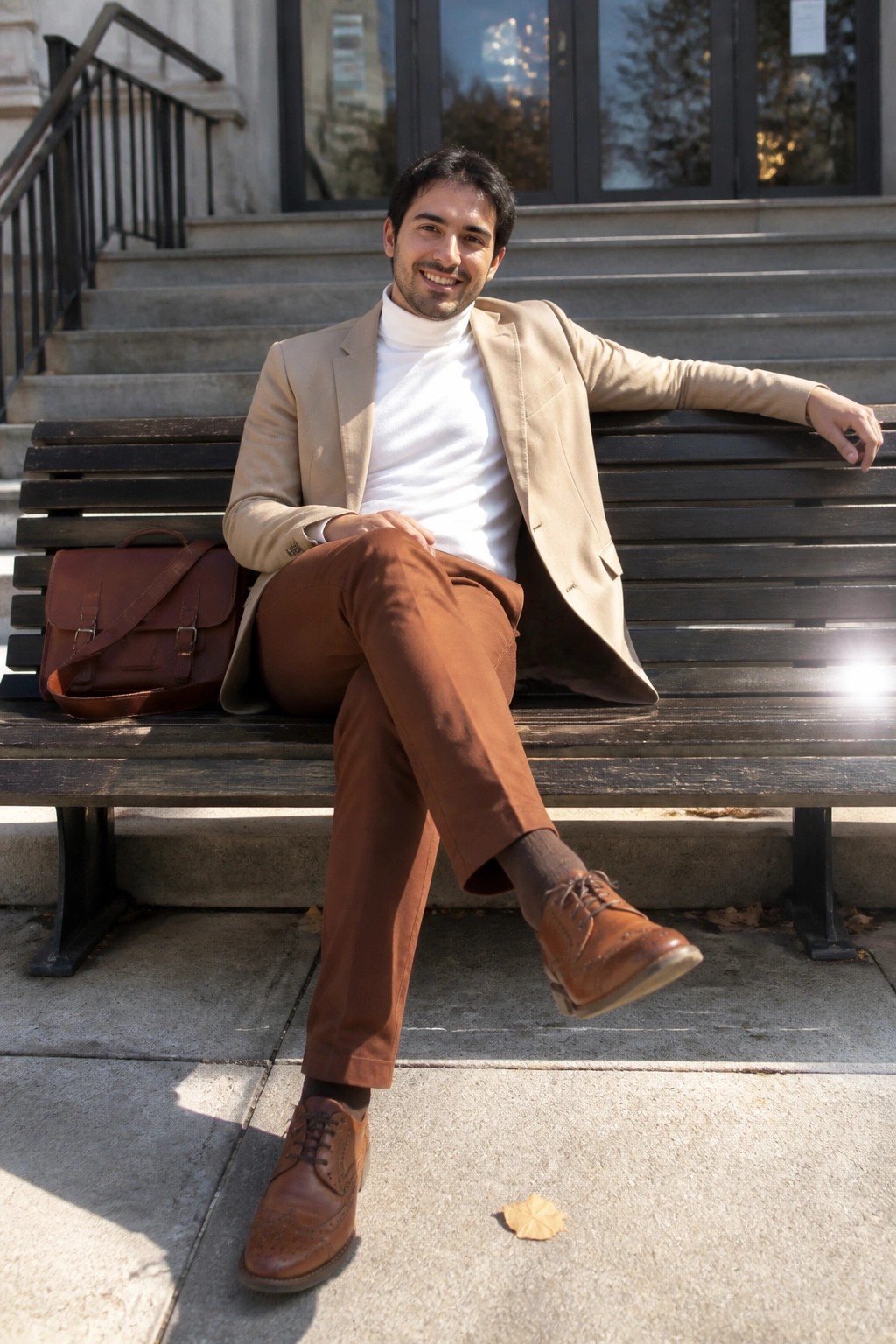 Man in beige blazer and white turtleneck sits on bench with brown bag