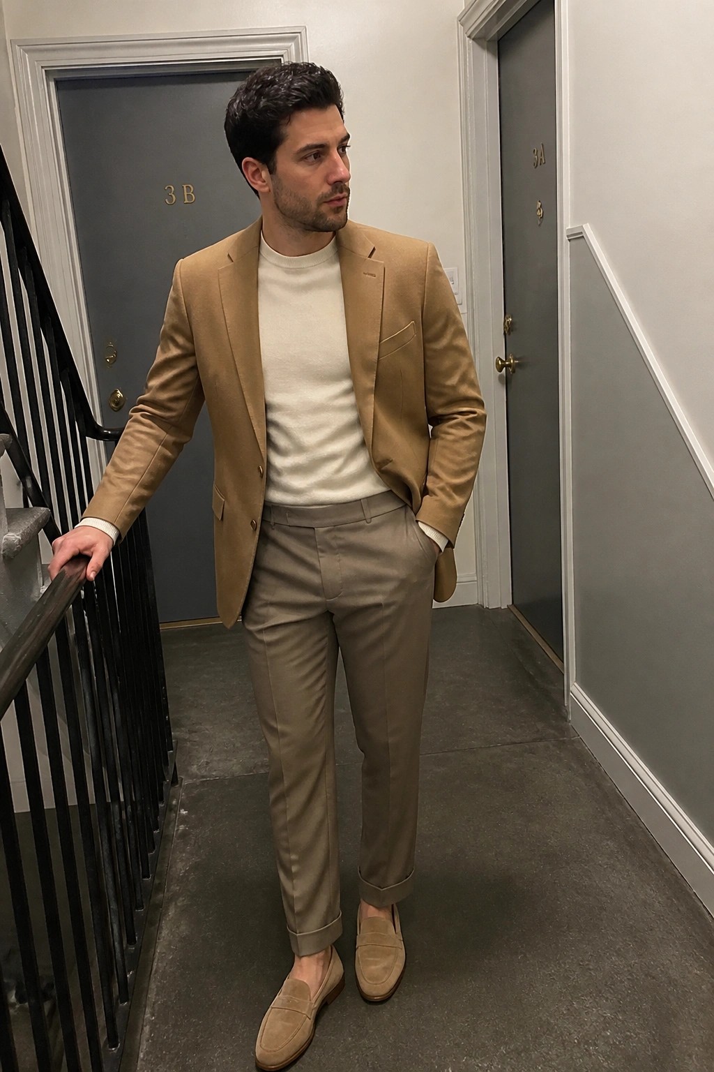 Man in tan blazer, cream sweater, and beige trousers stands in hallway.
