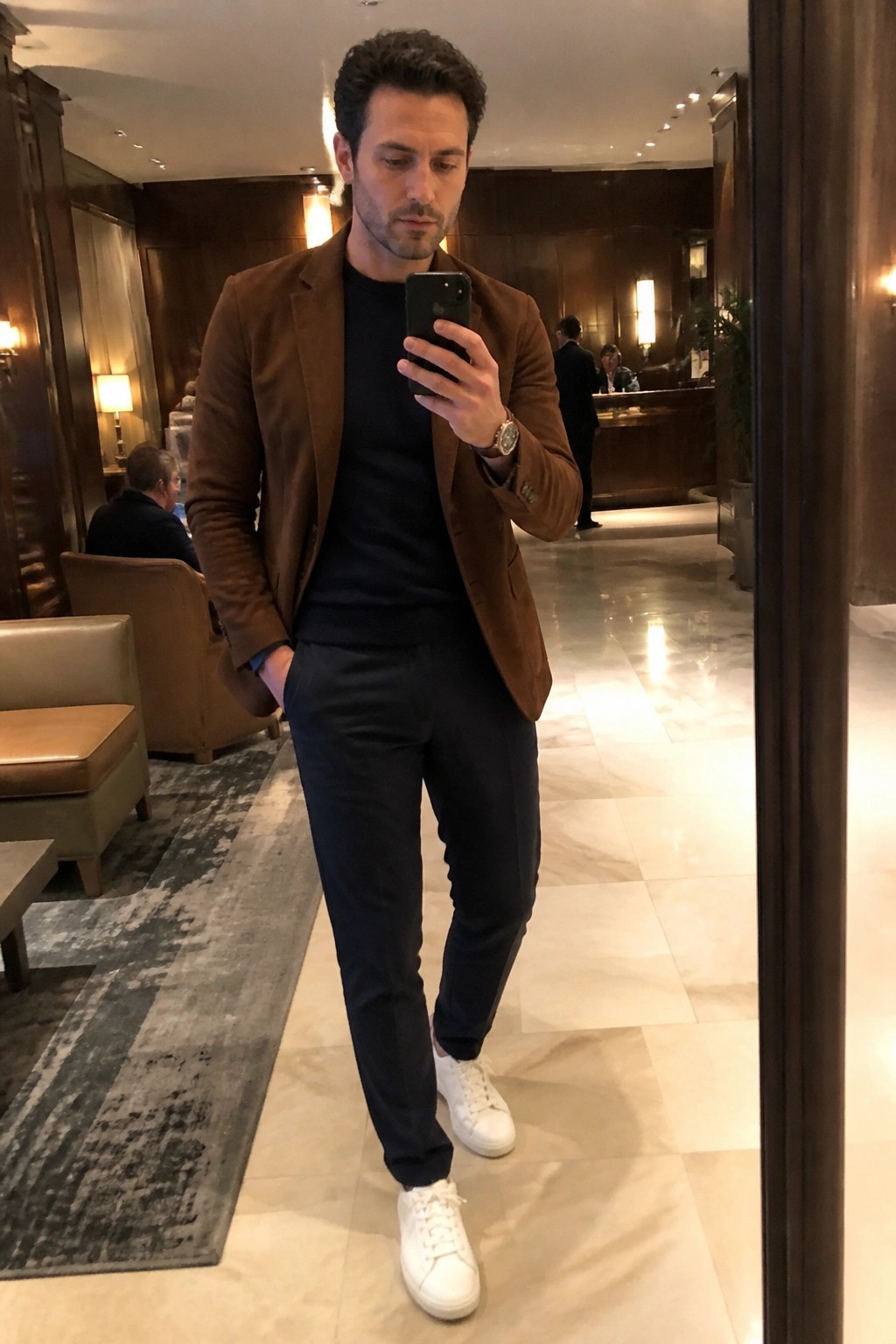 Man in brown blazer, black shirt, and white sneakers taking mirror selfie in lobby