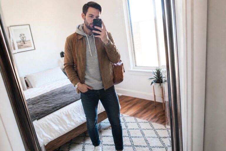 Man in brown suede jacket over gray hoodie taking mirror selfie in bedroom