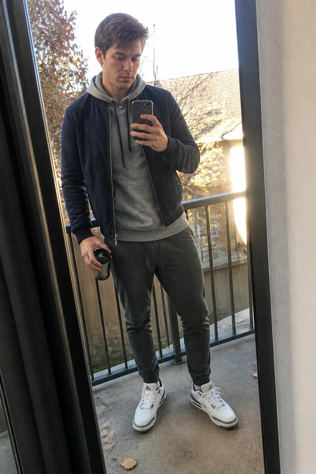 Man in navy bomber jacket and grey hoodie takes balcony mirror selfie with phone and mug