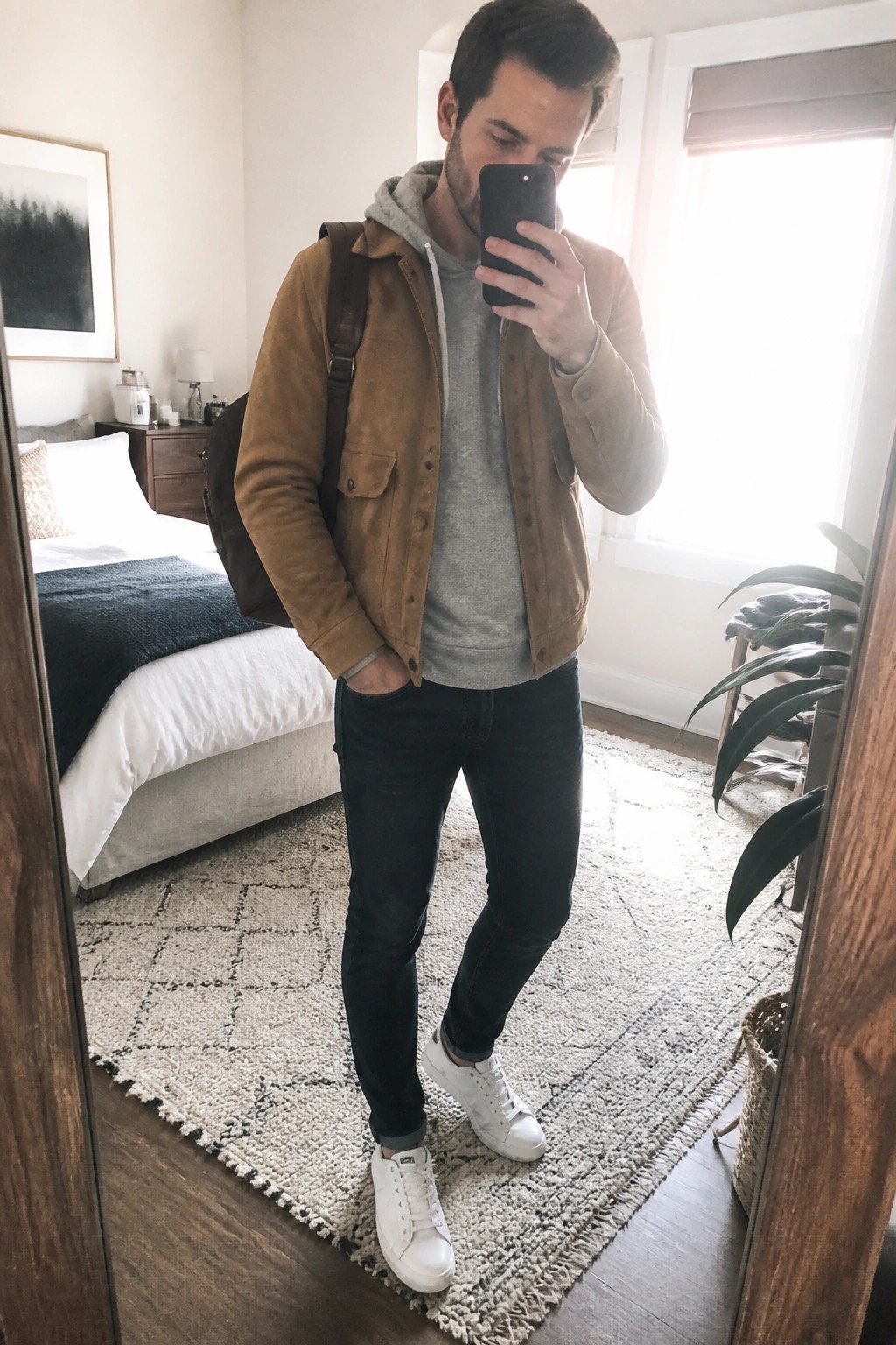Man in brown suede jacket over gray hoodie taking mirror selfie in bedroom