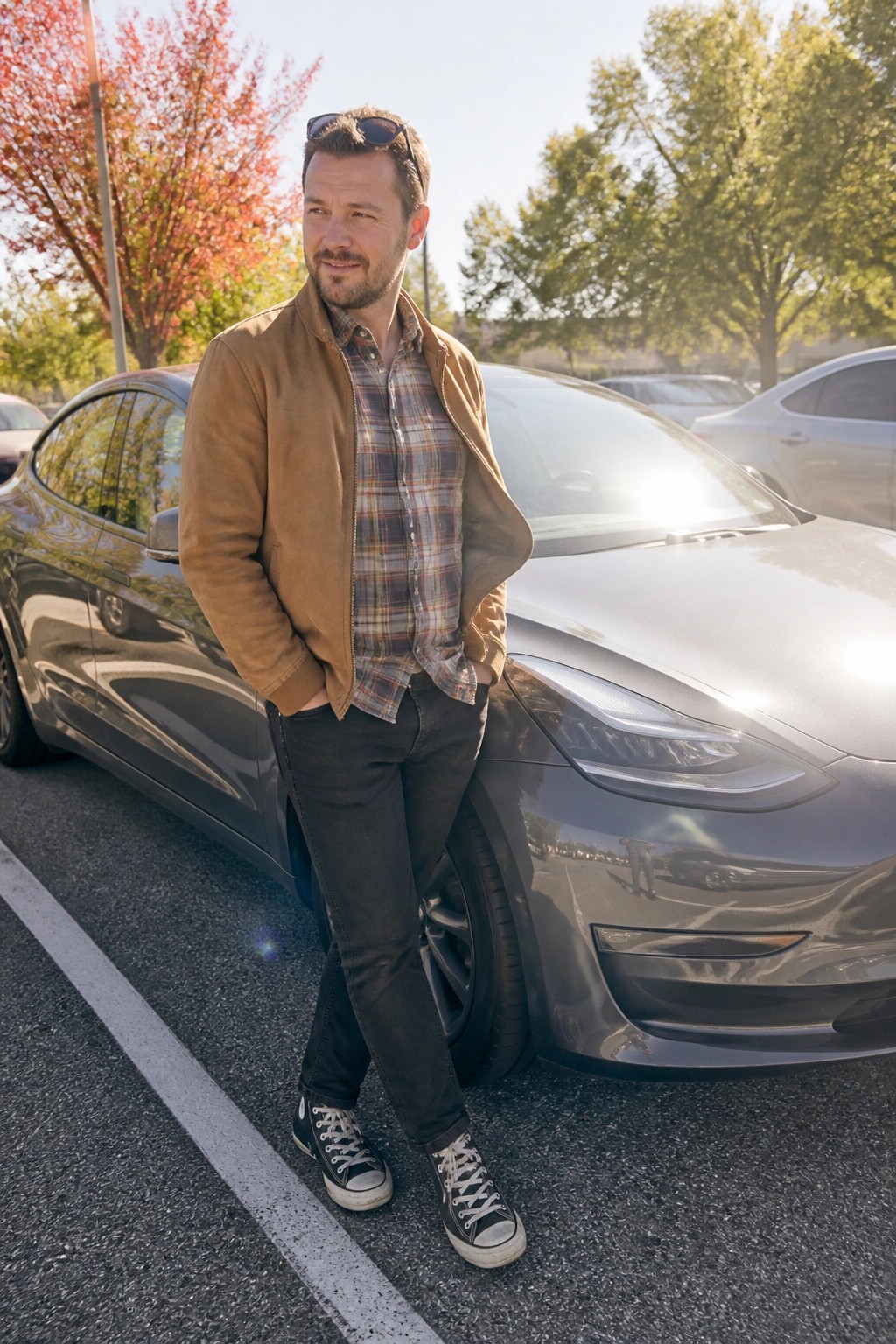 Man in brown jacket and plaid shirt leaning on dark gray Tesla in lot