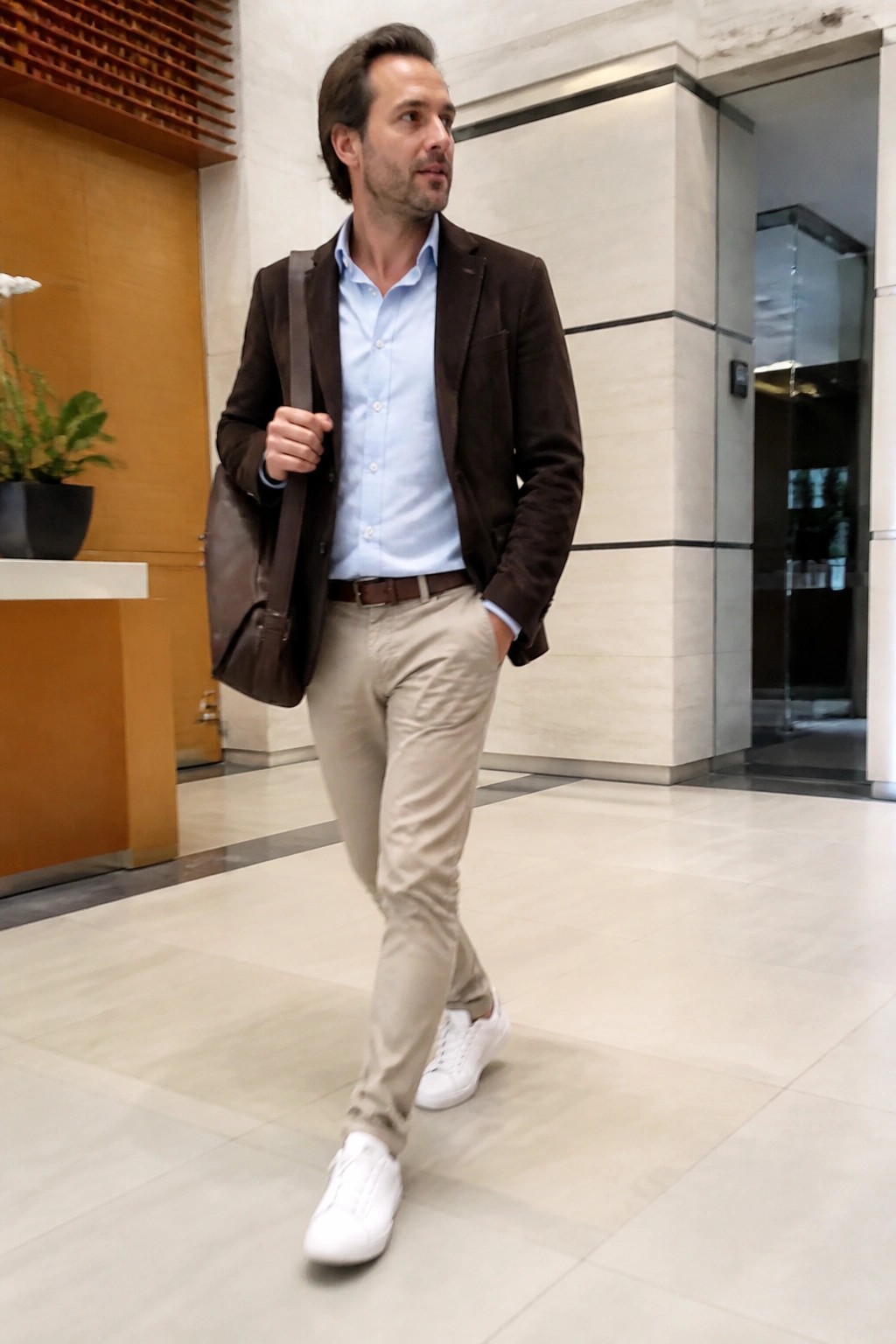 Man in brown jacket, blue shirt, beige pants carrying leather bag in modern lobby