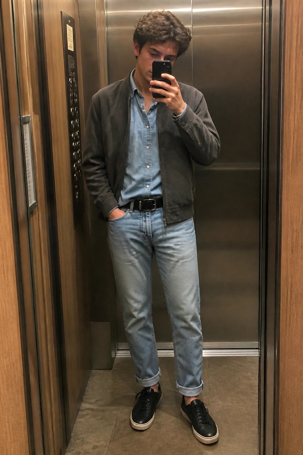 Young man in gray bomber jacket and jeans taking mirror selfie in elevator