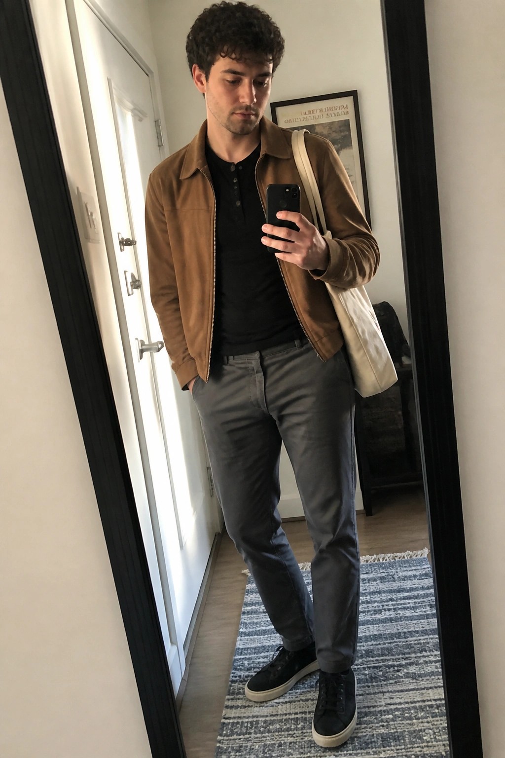 Man in brown jacket and gray pants takes mirror selfie with white tote bag