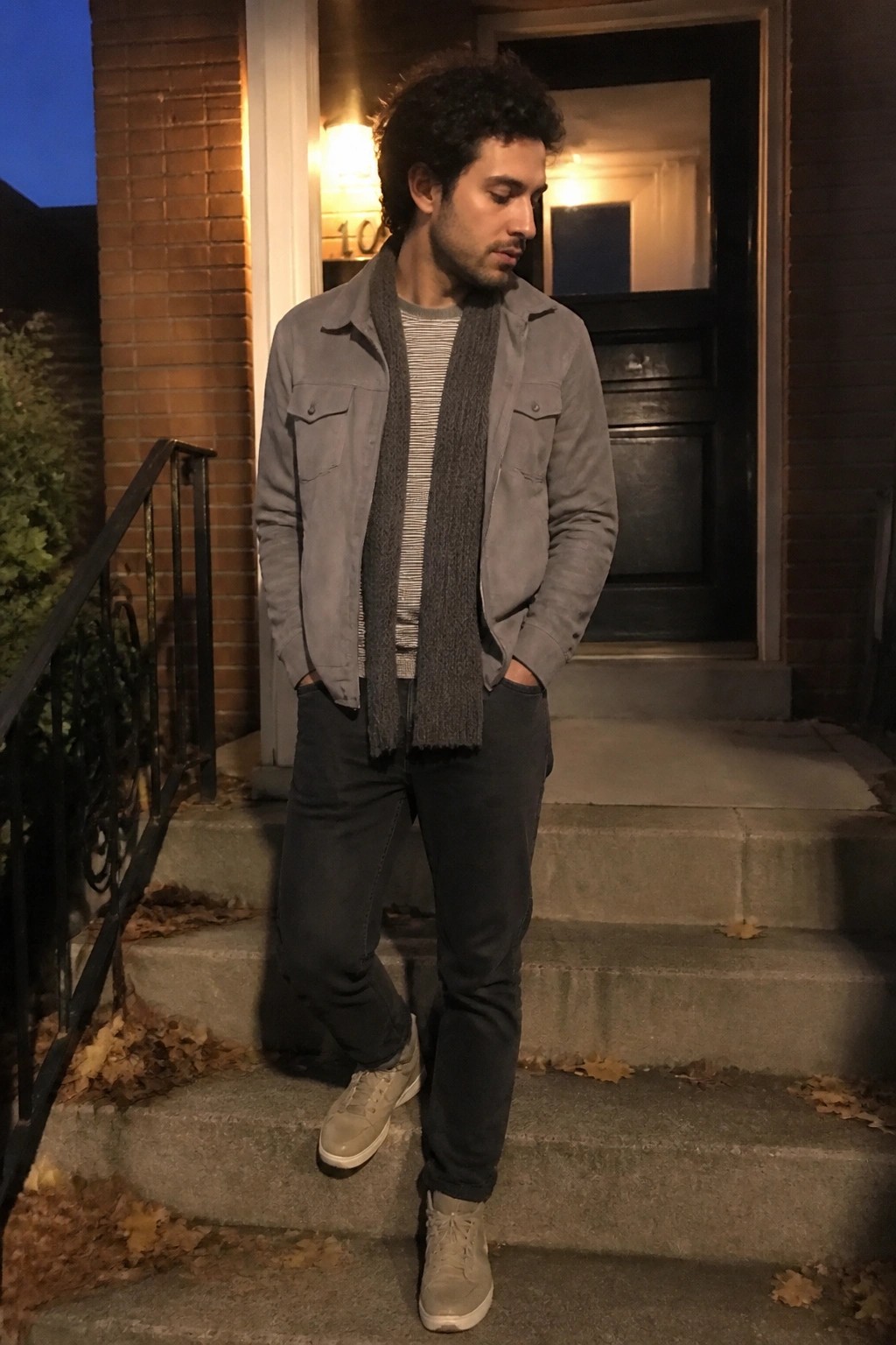 Man in gray suede jacket and scarf stands on brick house steps at dusk.