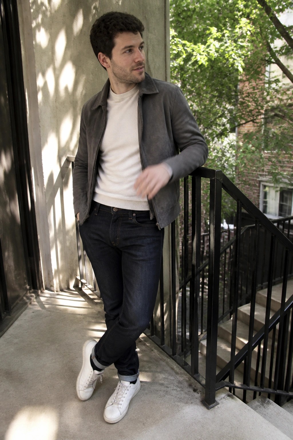 Man in gray suede jacket and white sweater leaning on outdoor railing