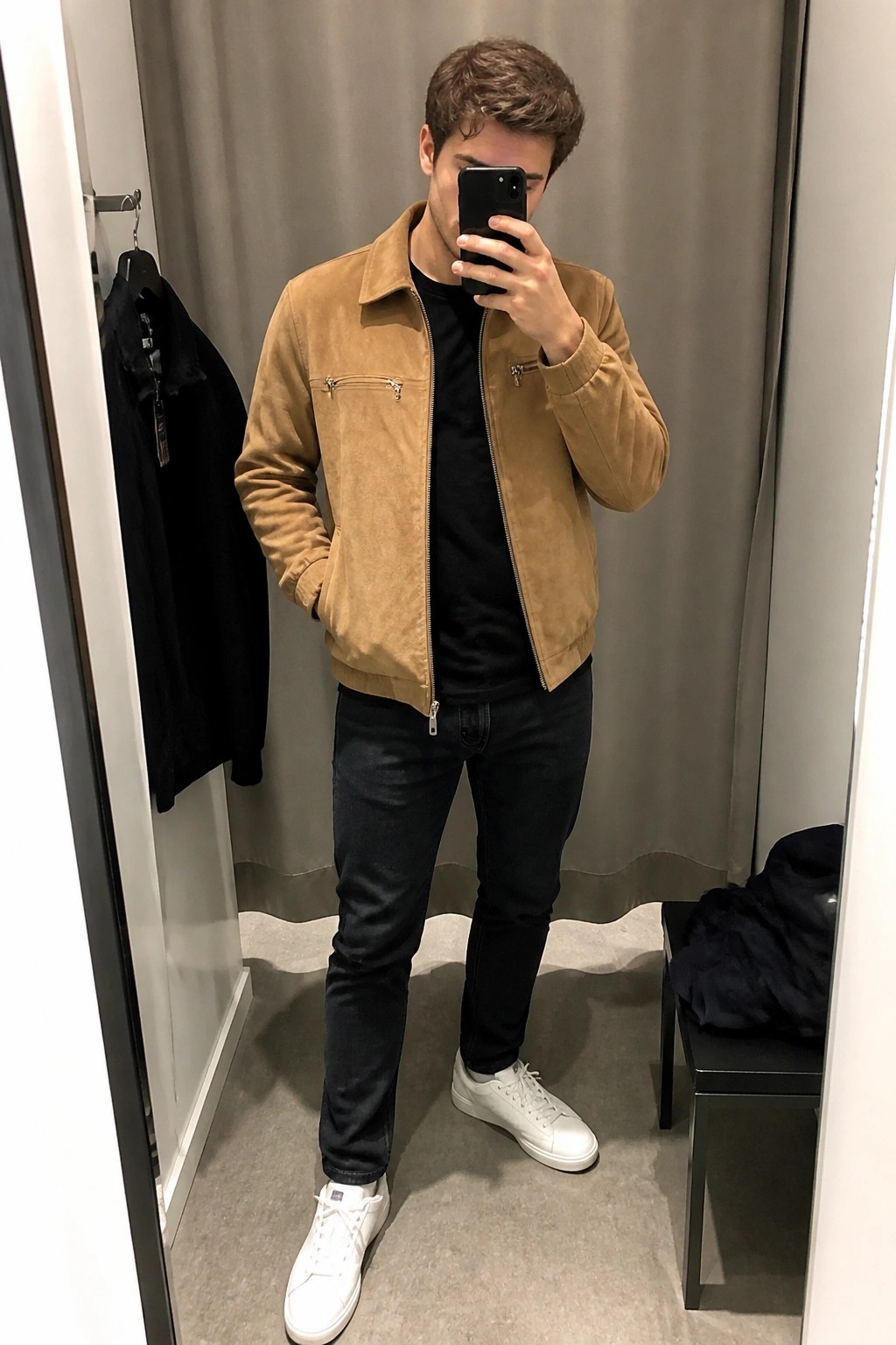 Man in tan suede jacket taking mirror selfie in fitting room wearing jeans and sneakers