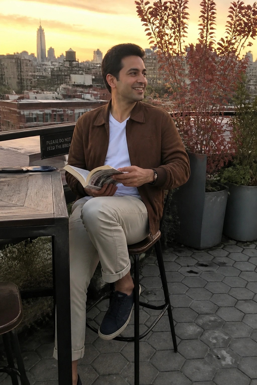 Man in brown jacket reads open book on rooftop overlooking city skyline at sunset