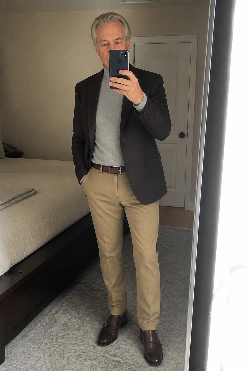 Man in dark blazer over gray turtleneck with khaki pants taking mirror selfie