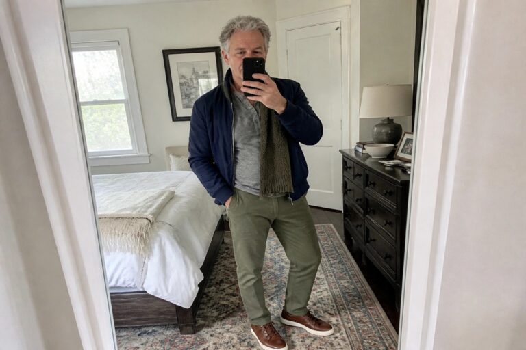 Gray-haired man in blue jacket, green pants, and knit scarf takes bedroom mirror selfie.