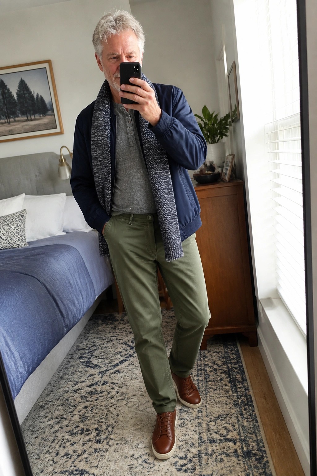 Gray-haired man in blue jacket, green pants, and knit scarf takes bedroom mirror selfie.