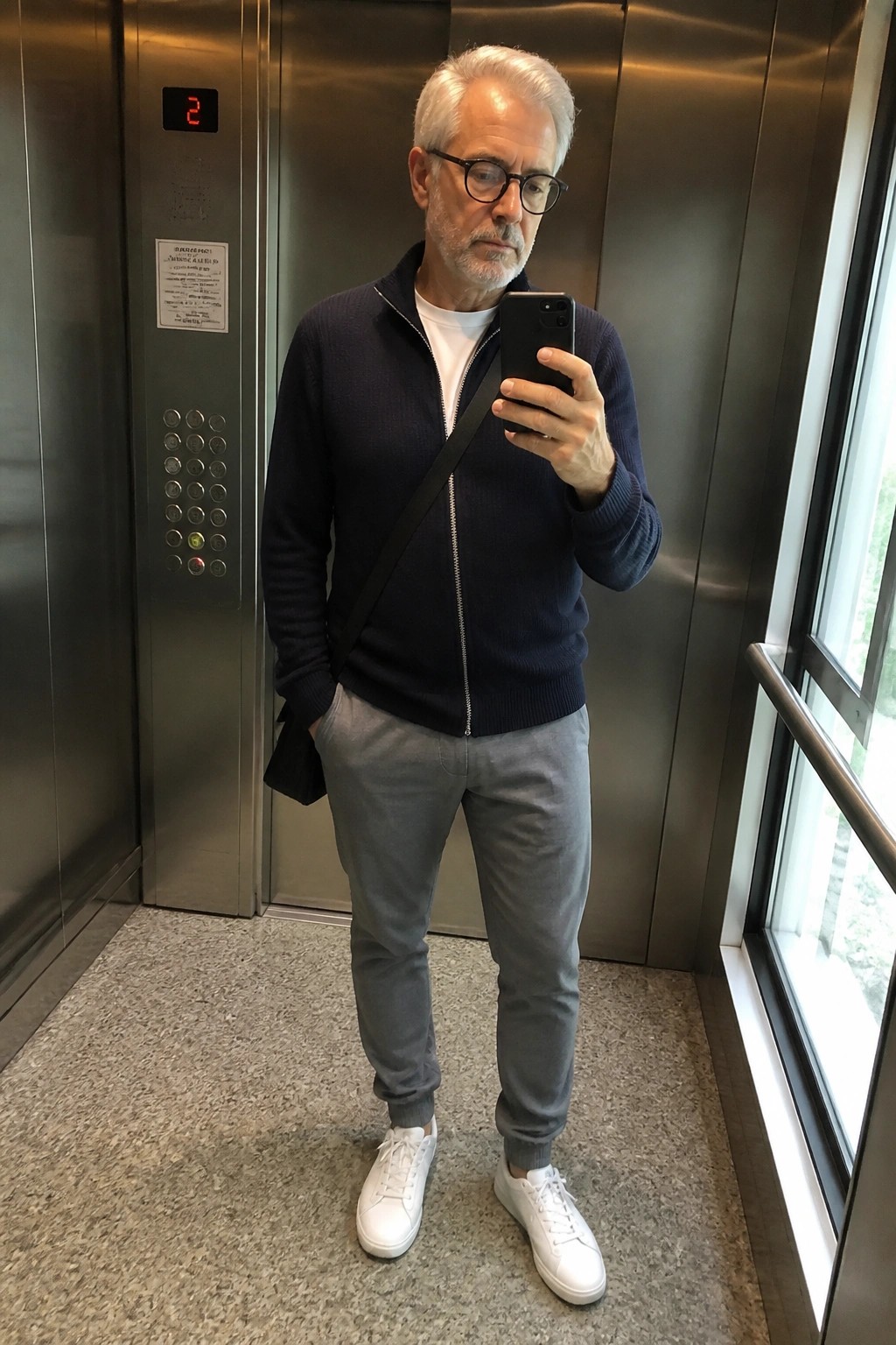 Gray-haired man in navy zip sweater takes phone selfie in elevator.
