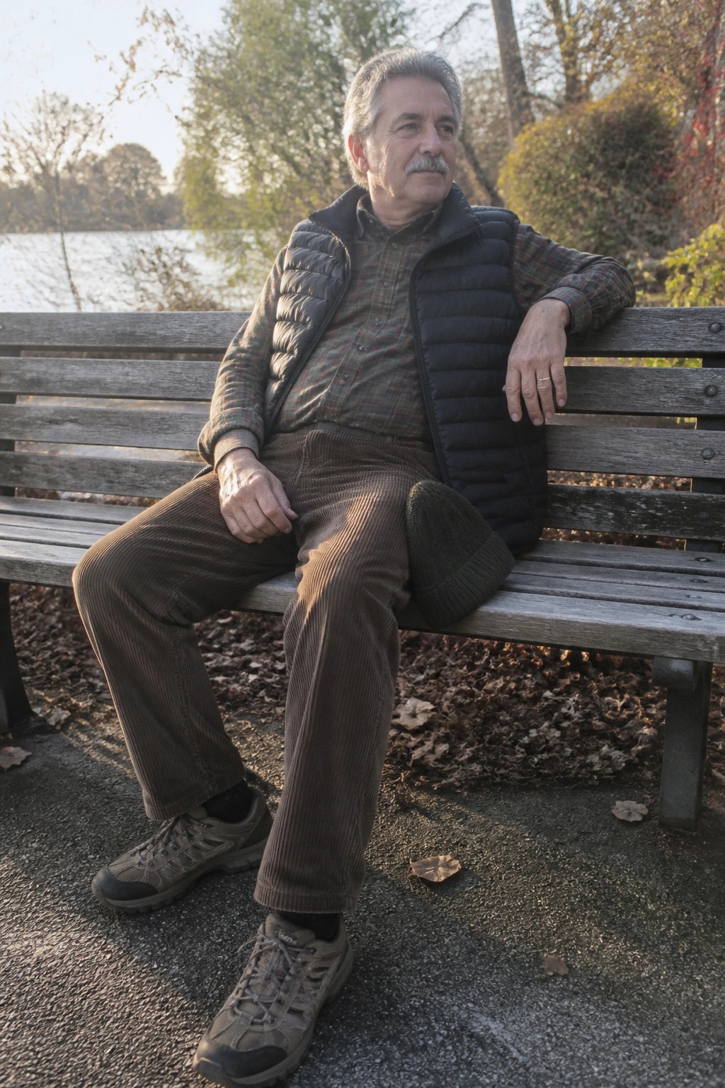 Older man with mustache sits on wooden bench by lake in autumn park wearing vest and corduroys.