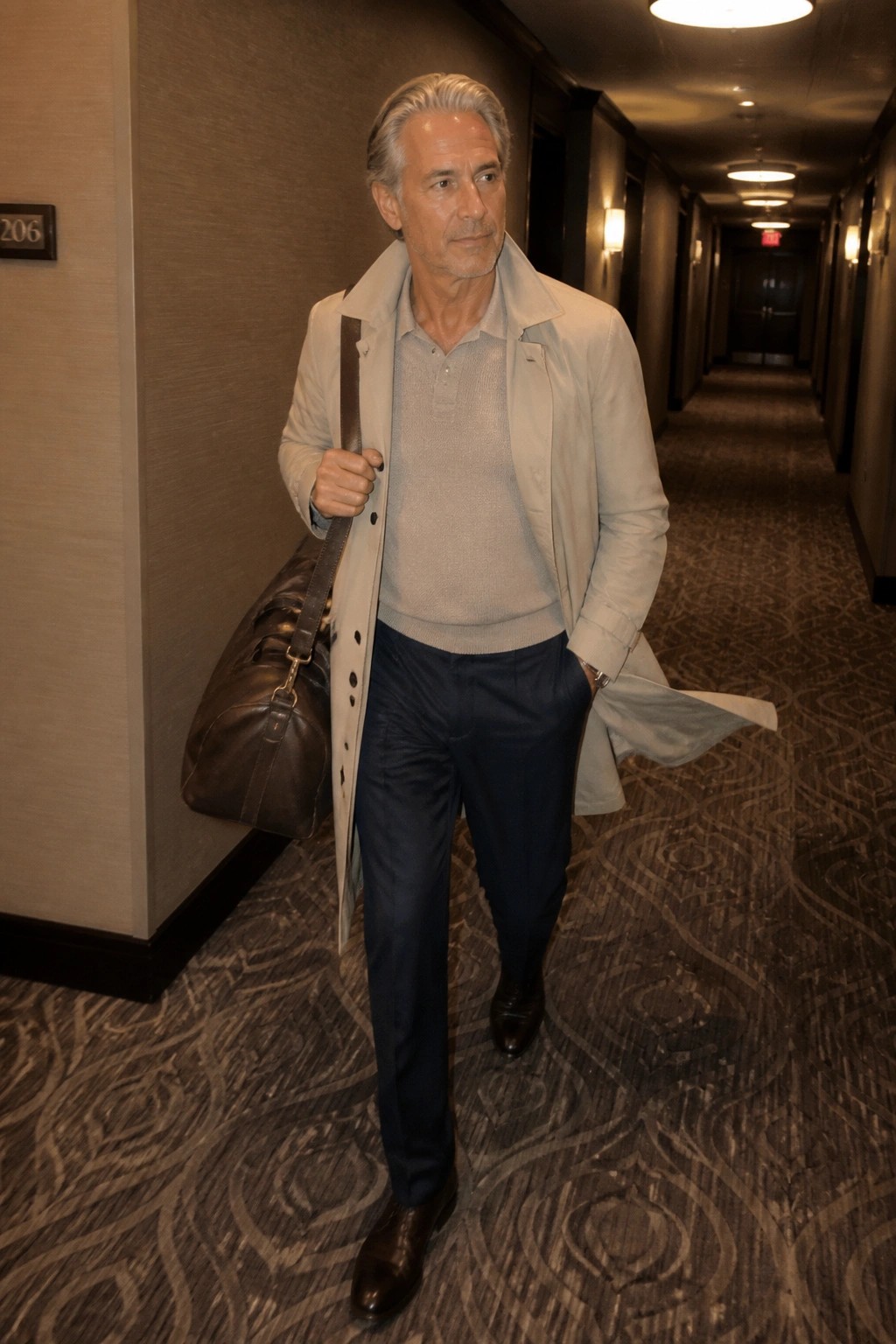Man in beige trench coat carries brown leather bag down hotel hallway