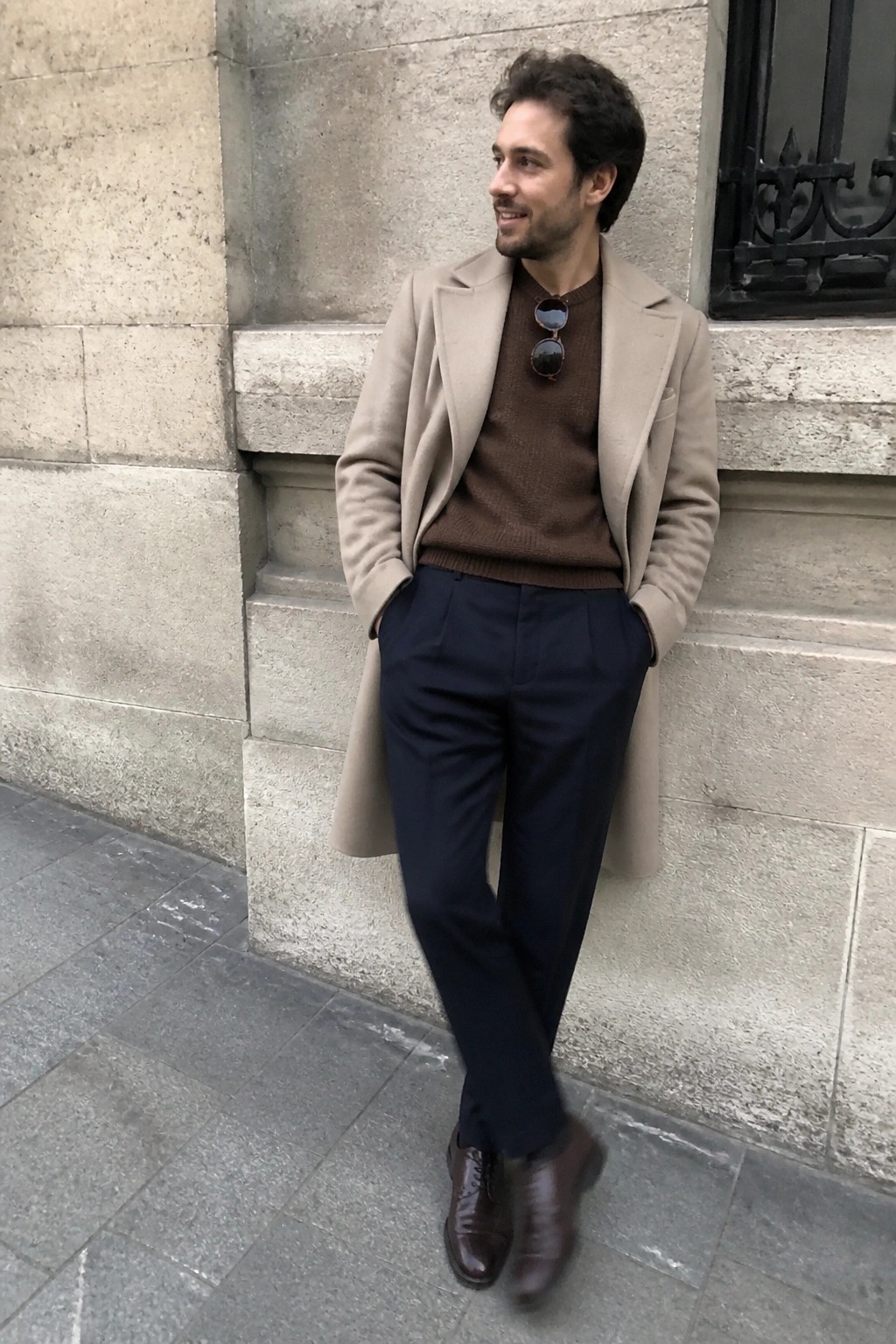Man in beige overcoat, brown sweater, navy trousers, brown shoes by stone wall